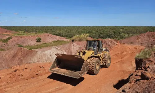Front-end loader driving along a red dirt road at a mining site with bushland in the background.